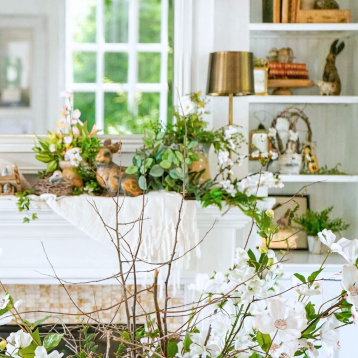 A bright, airy room with white shelves and a mantle decorated with greenery, flowers, animal figurines, and vintage Easter decorating ideas. A gold lamp, books, and more floral arrangements add a fresh, spring-like feel.