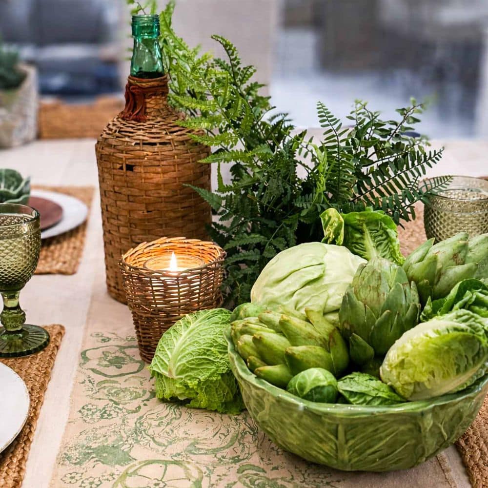 A table decorated with green glassware, a woven bottle, a candle in a wicker holder, ferns, and a bowl filled with lettuce, cabbage, and artichokes, creating a fresh, rustic centerpiece.