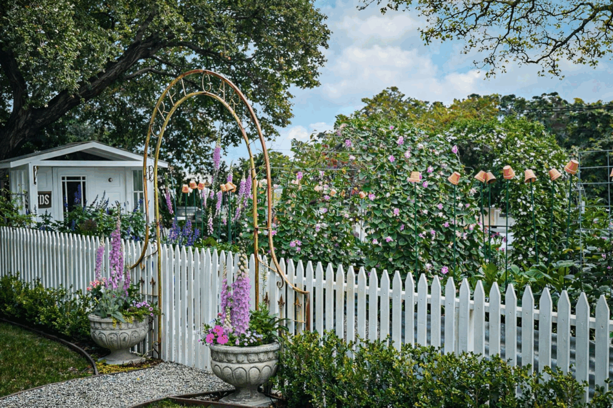 A charming garden with blooming flowers, a white picket fence, and a metal archway. Two large flower pots flank the entrance, and a small white house is visible in the background under leafy trees.