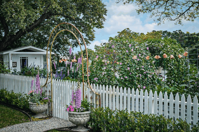 A charming garden with blooming flowers, a white picket fence, and a metal archway. Two large flower pots flank the entrance, and a small white house is visible in the background under leafy trees.