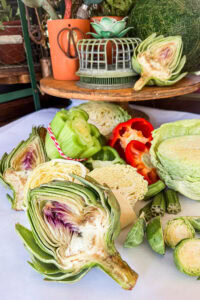 A colorful assortment of fresh vegetables, perfect for vegetable block printing, includes halved artichokes, celery, red bell pepper, cabbage, okra, and Brussels sprouts on a white surface with a wooden shelf and decorative items in the background.