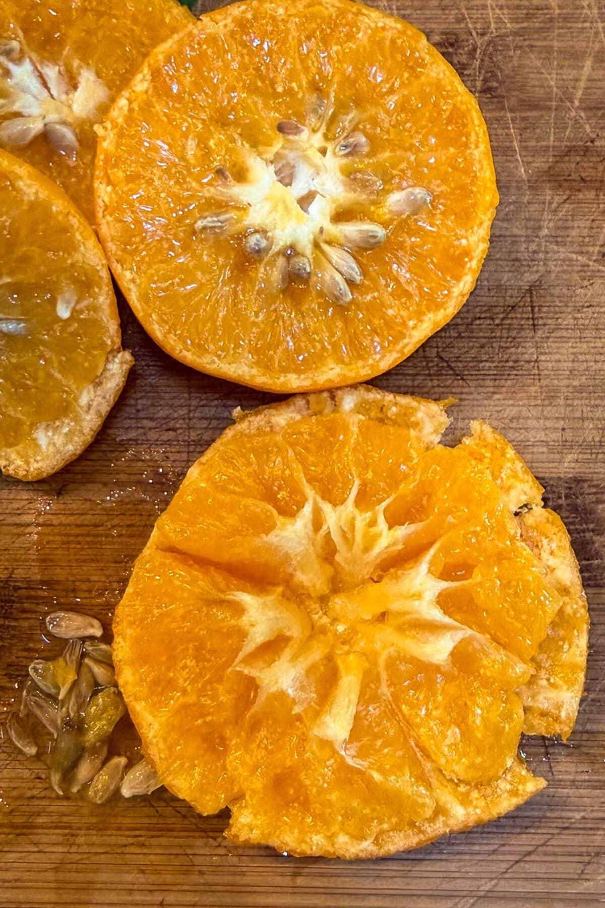 Two halves of an orange rest on a wooden cutting board. One half is tidy and shows its seeds, while the other is roughly cut and segmented, with some seeds—and traces of tangerine puree—spilled nearby.