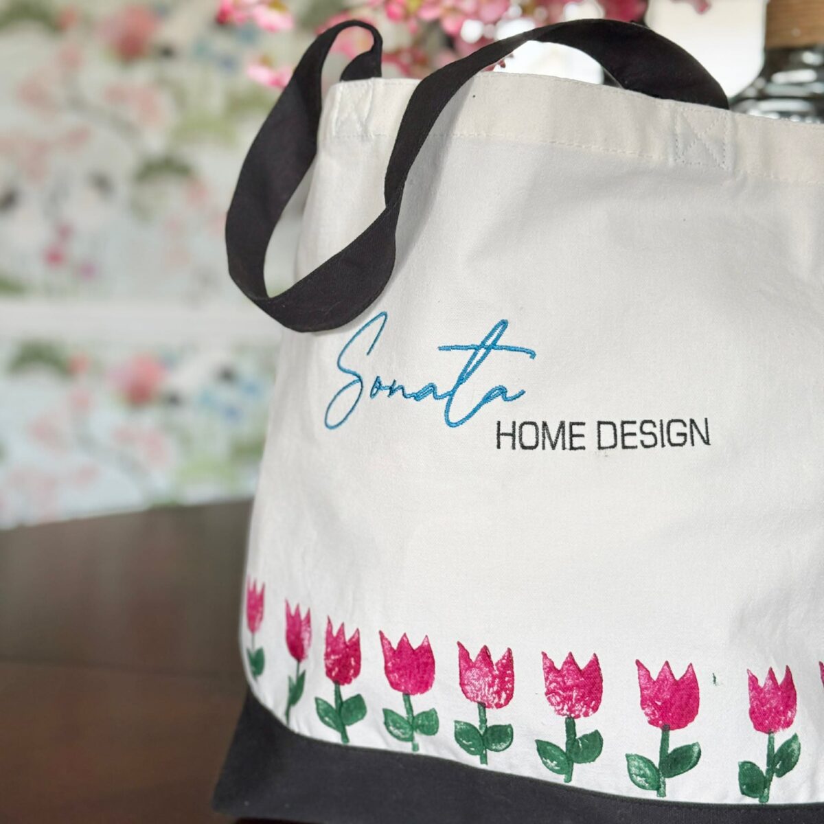 A white tote bag with black handles displays "Sonata HOME DESIGN" in blue and black text. Pink tulip patterns line the bottom. Blurred flowers and wallpaper are visible in the background.