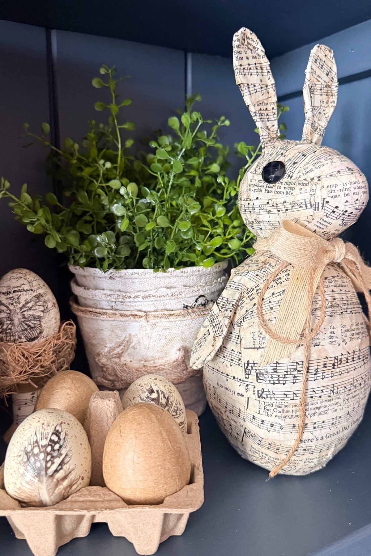 A Paper Mache Easter Bunny covered in sheet music sits next to a potted green plant and several eggs, some in a cardboard holder and others decorated with vintage designs—all arranged on a dark shelf.