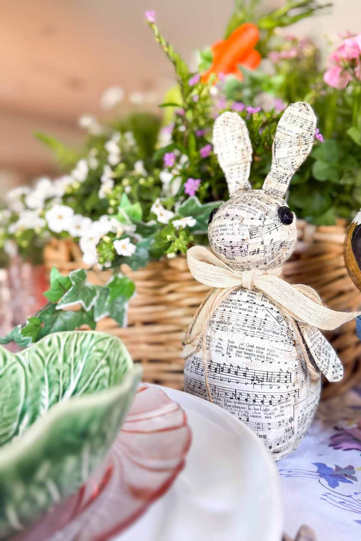 A decorative Paper Mache Easter Bunny made from sheet music sits beside a plate, with a wicker basket of colorful flowers in the background, creating a cheerful and festive table setting.