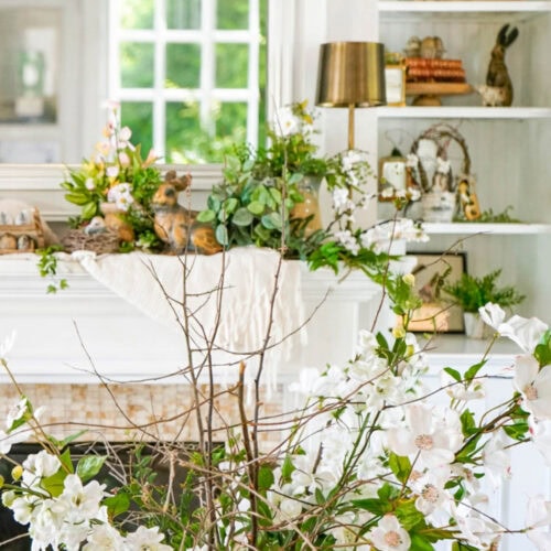 A bright, cozy living room with white shelves, books, and spring decor. A vase of blooming white flowers sits in front of a mantel decorated with greenery, bunnies, and a mirror—perfect for vintage Easter decorating ideas.
