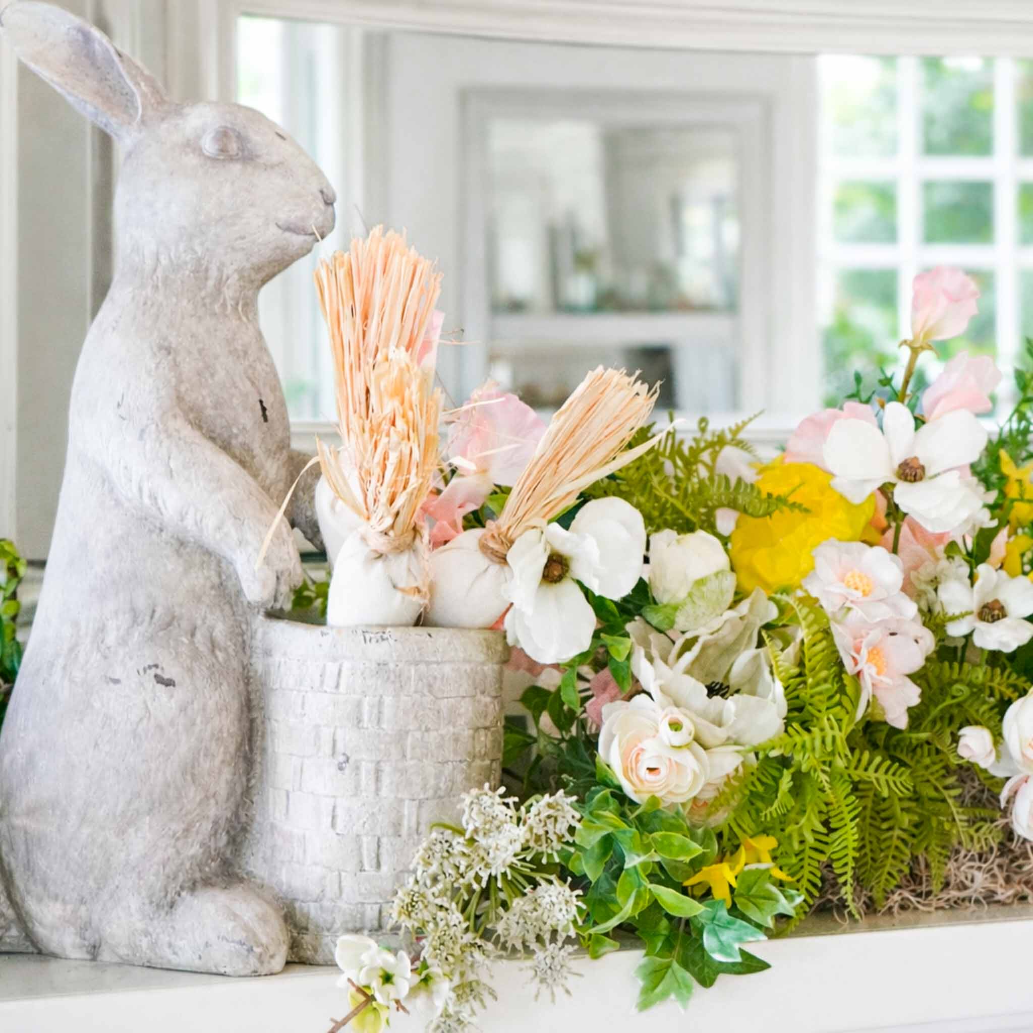 A stone bunny statue stands beside a white basket filled with pink and white artificial flowers, greenery, and yellow accents—simple Easter decor for spring decorating ideas on a white mantel in front of a mirror.