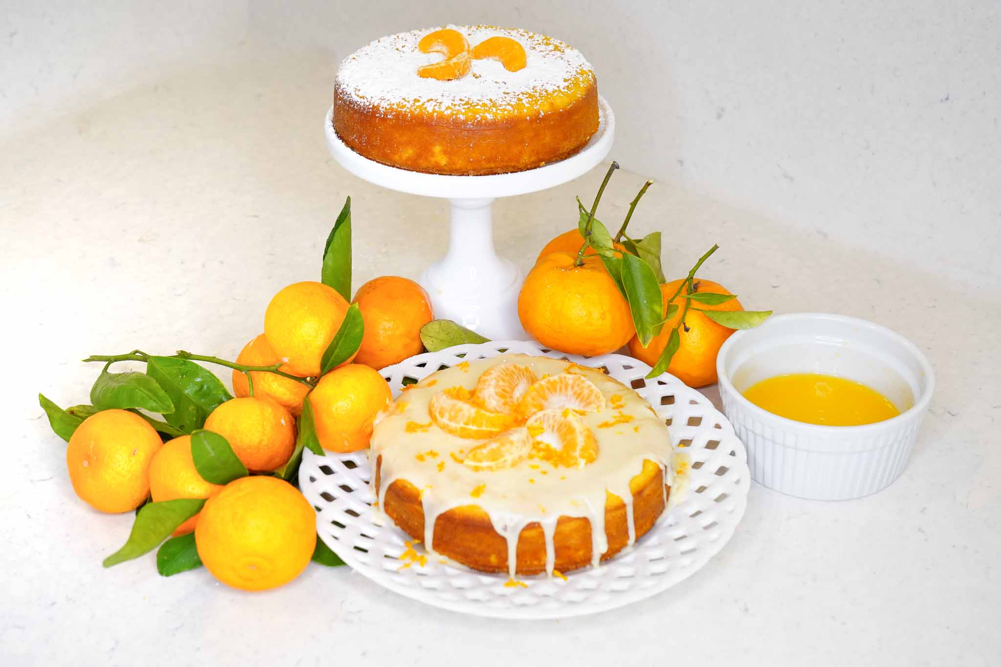 Two cakes are displayed: one on a stand dusted with powdered sugar and topped with mandarin slices, and another glazed with icing and tangerine puree alongside mandarin segments. Fresh mandarins and a bowl of sauce sit beside them.