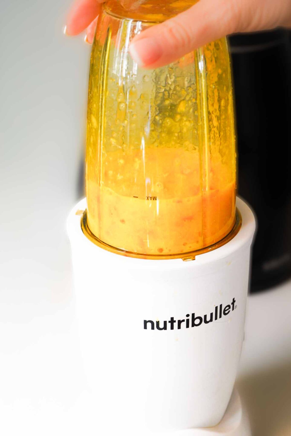 A hand holds a blending cup filled with homemade tangerine puree in a white Nutribullet blender, blending ingredients.
