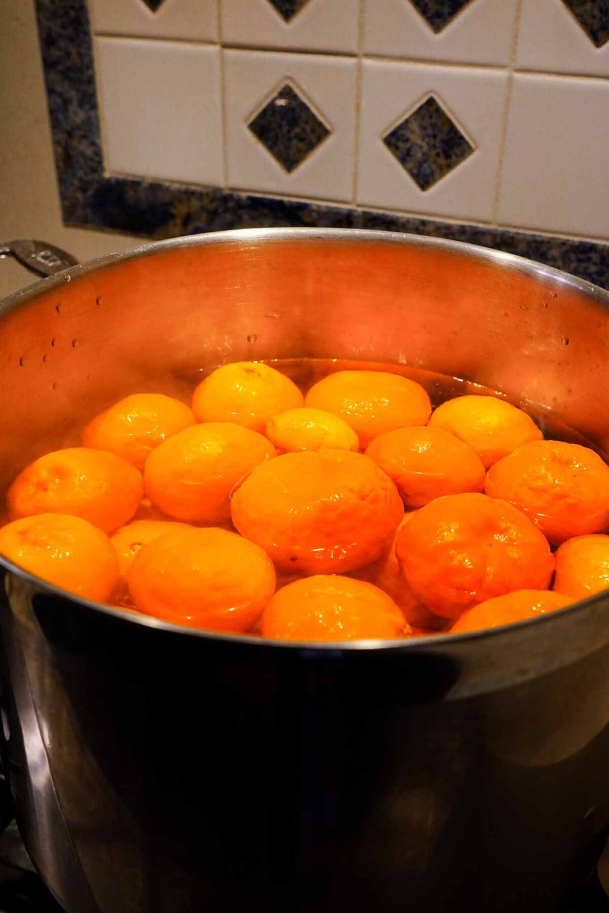 A large pot filled with whole oranges boiling in water on a stovetop, with a tiled backsplash visible in the background.