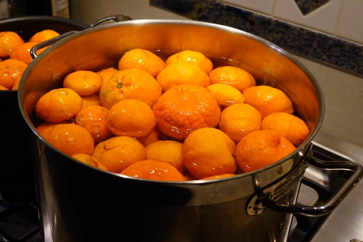 A stainless steel pot filled with whole oranges submerged in water, sitting on a stovetop, with another pot partially visible in the background.
