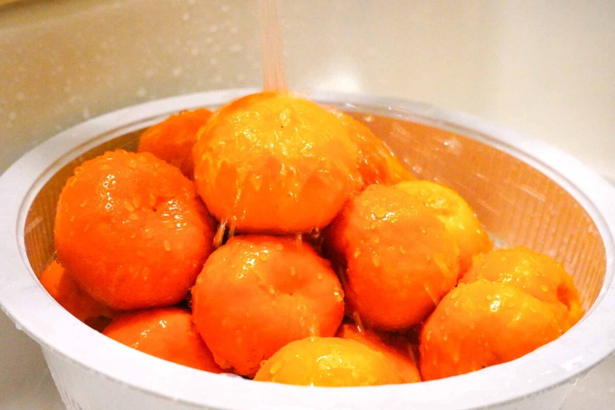 A close-up of peeled tomatoes piled in a white colander under running water, with droplets glistening on their surfaces.