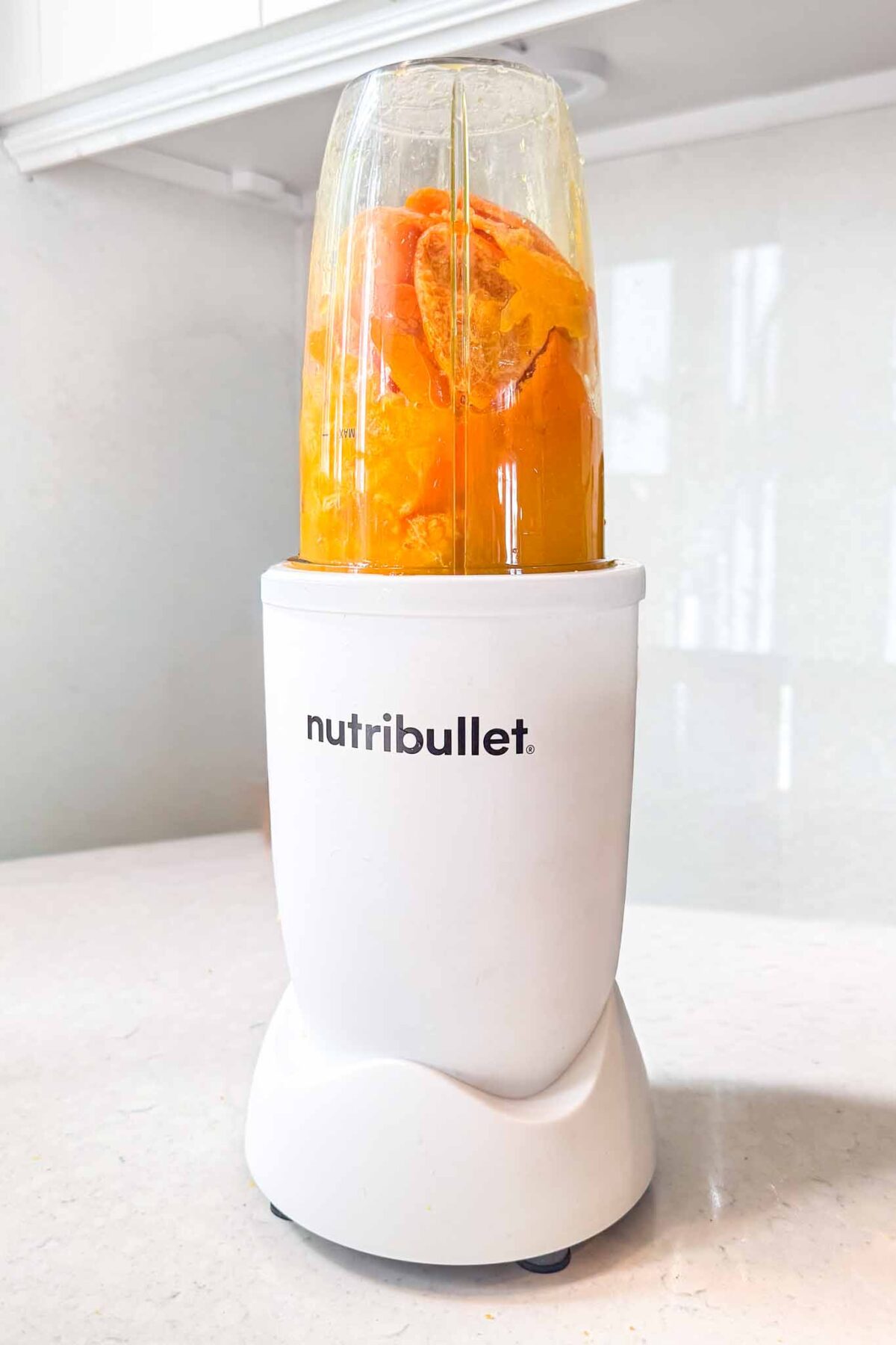 A white Nutribullet blender filled with orange fruit pieces sits on a light kitchen counter, ready to blend.