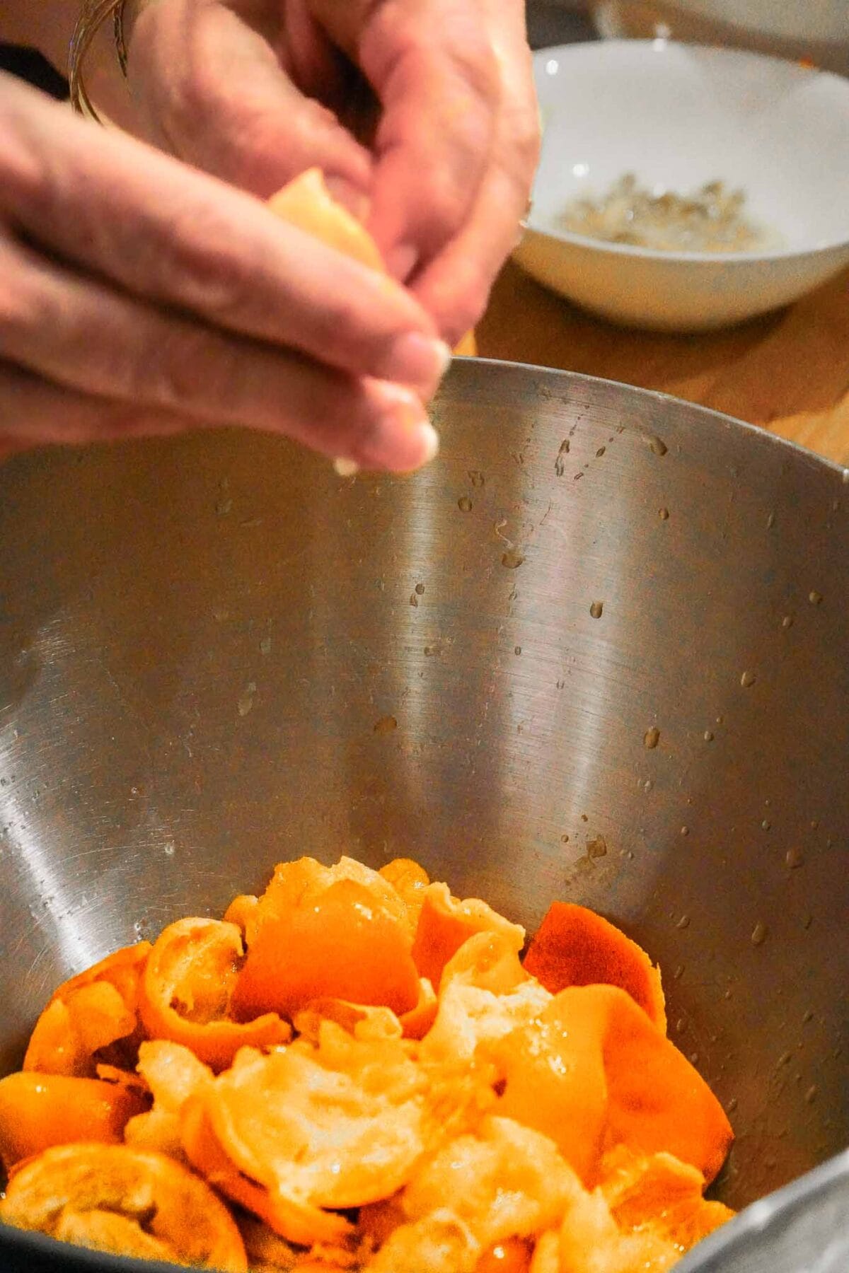 A person squeezes citrus fruit halves over a metal bowl filled with squeezed orange and lemon peels, with a white dish in the background.
