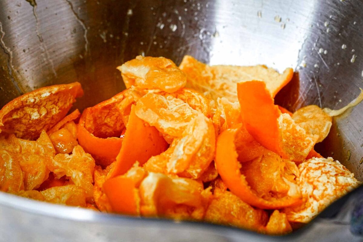 A close-up of orange peels and segments in a stainless steel bowl, showing the vibrant color and texture of the fruit and its rind.