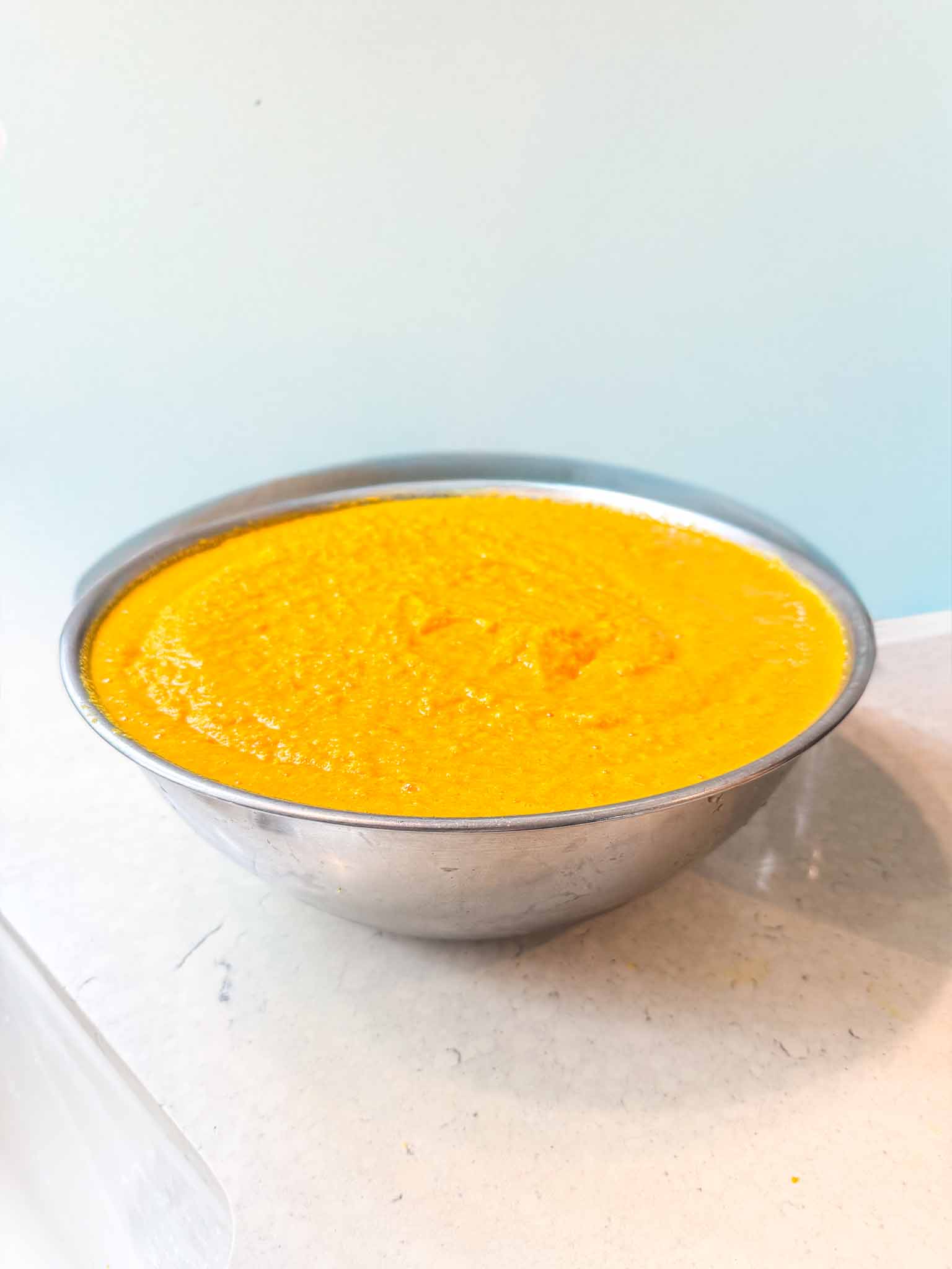 A large metal mixing bowl filled to the brim with a smooth, bright yellow-orange puree sits on a light-colored countertop.