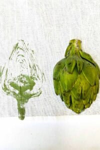 A fresh green artichoke sits on white fabric next to a green paint print of the artichoke's cross-section, showcasing a creative vegetable block printing art project.