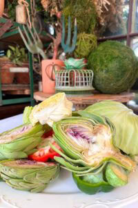 Halved artichokes, cabbage, and red bell pepper are arranged on a white plate with gardening tools, potted plants, and a ball of moss in the background. The setting appears rustic and natural.