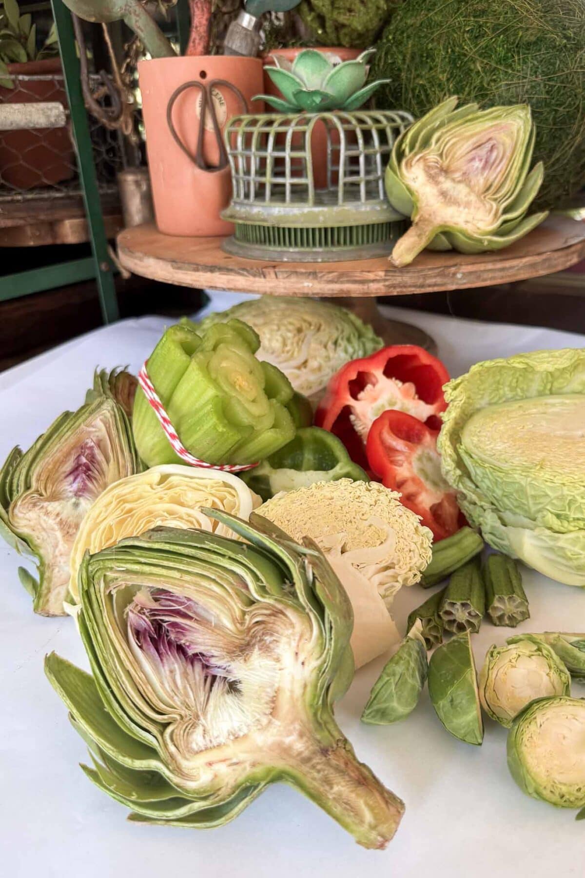 A variety of fresh, sliced vegetables including artichokes, celery, cabbage, bell pepper, and okra are arranged on a white surface with potted plants and decorative items in the background.
