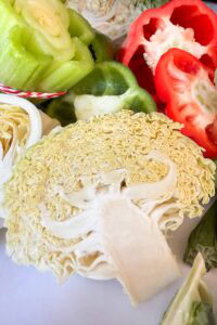 A close-up of a sliced cabbage revealing intricate patterns, surrounded by sliced green and red bell peppers and other vegetable pieces on a white surface.