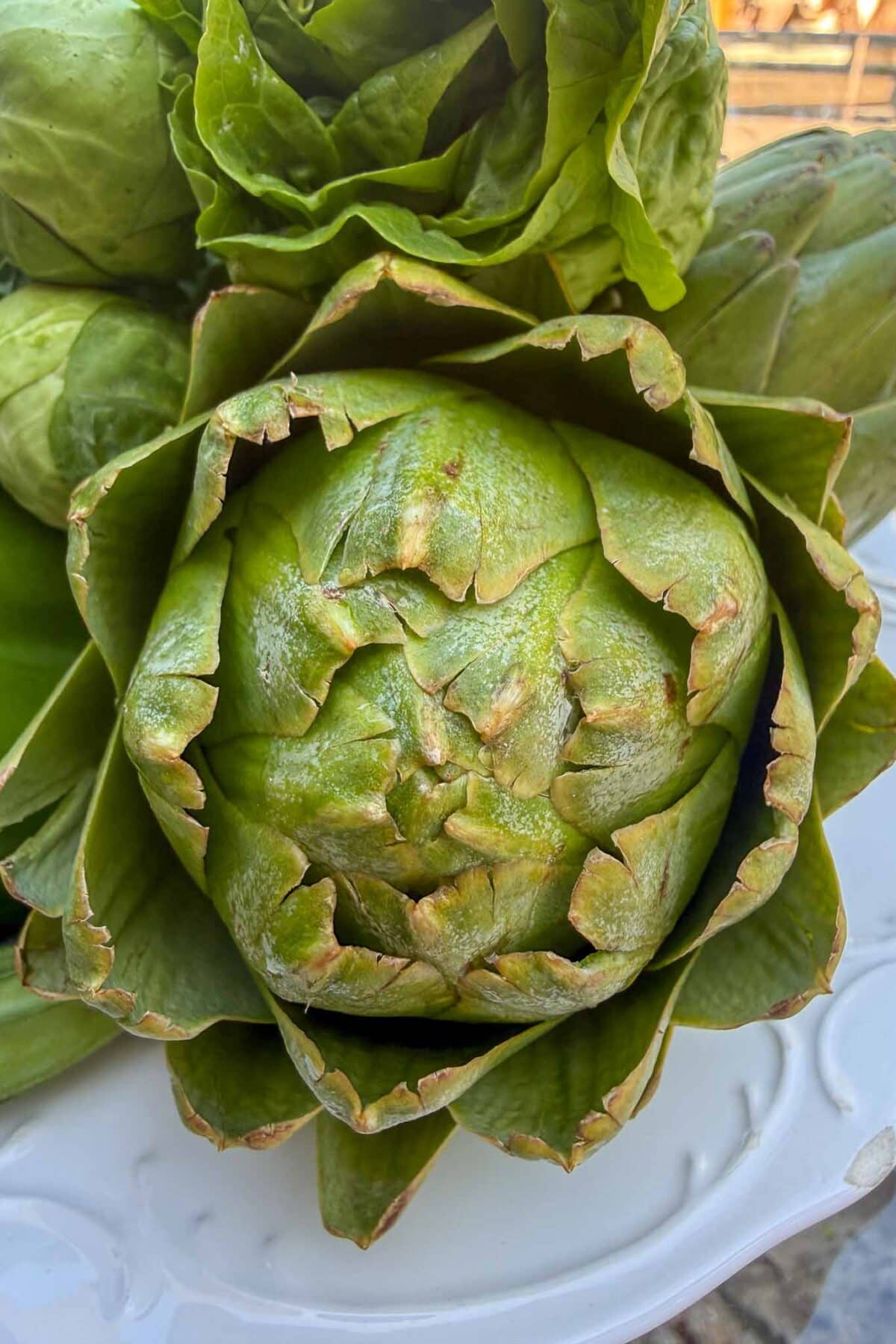 Close-up of a fresh green artichoke with tightly packed leaves, some of which are slightly brown at the tips, displayed on a white surface.