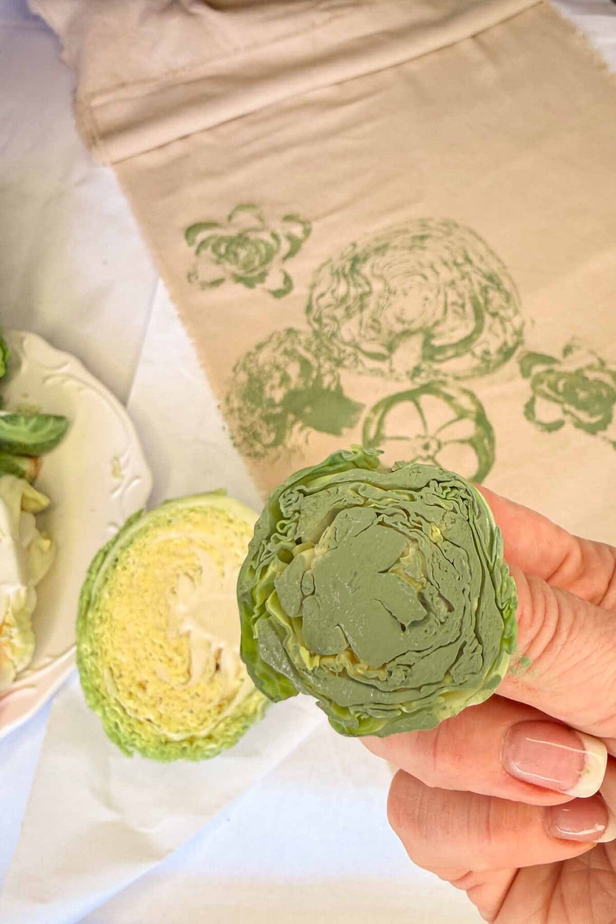 A hand holds a halved cabbage painted green, ready to use as a stamp. In the background, a fabric piece shows cabbage and other vegetable prints in green ink.