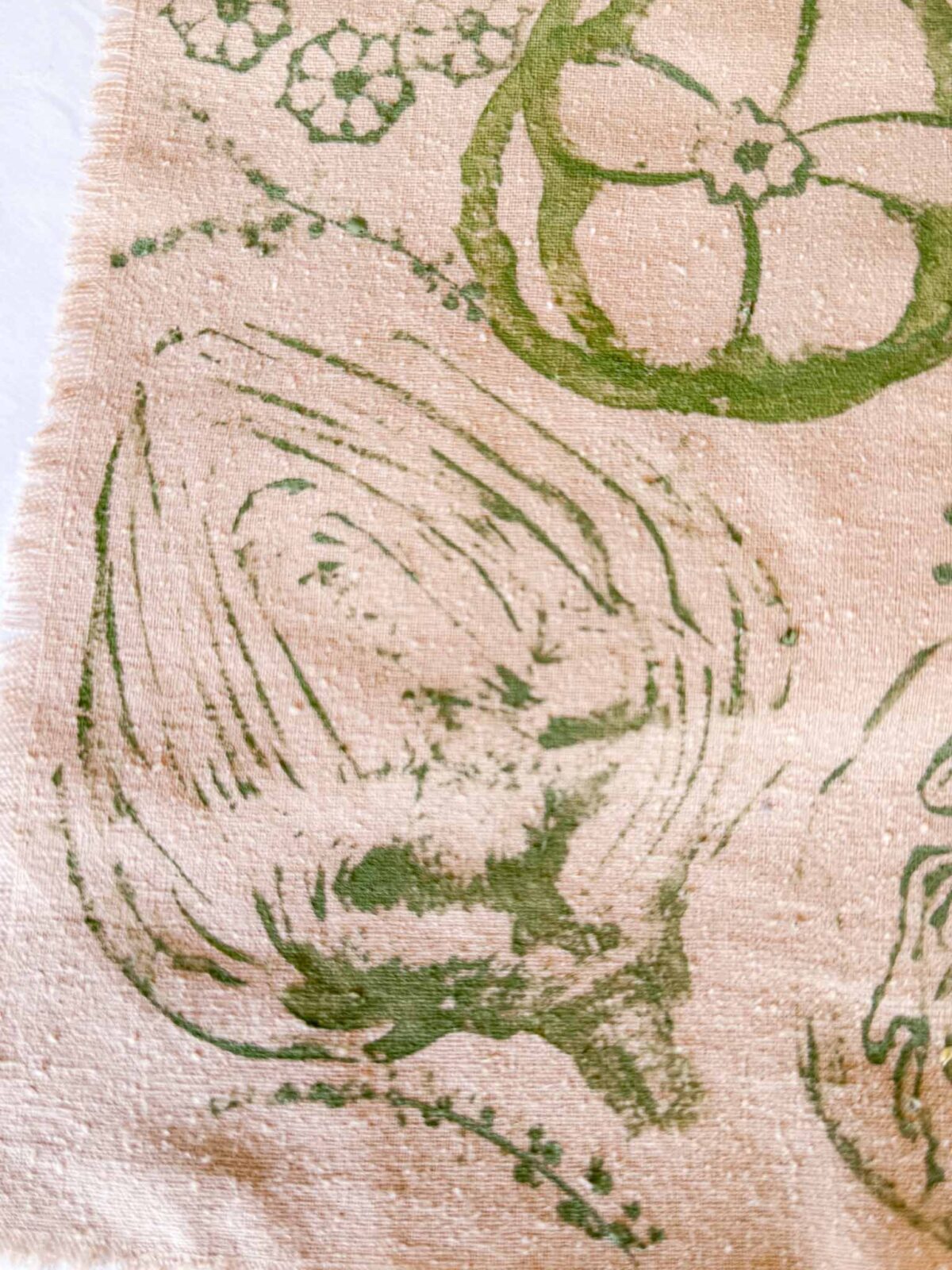 A beige fabric with green botanical prints, including an artichoke and a cross-section of a bell pepper. The fabric has a textured surface and a frayed edge on the left side.