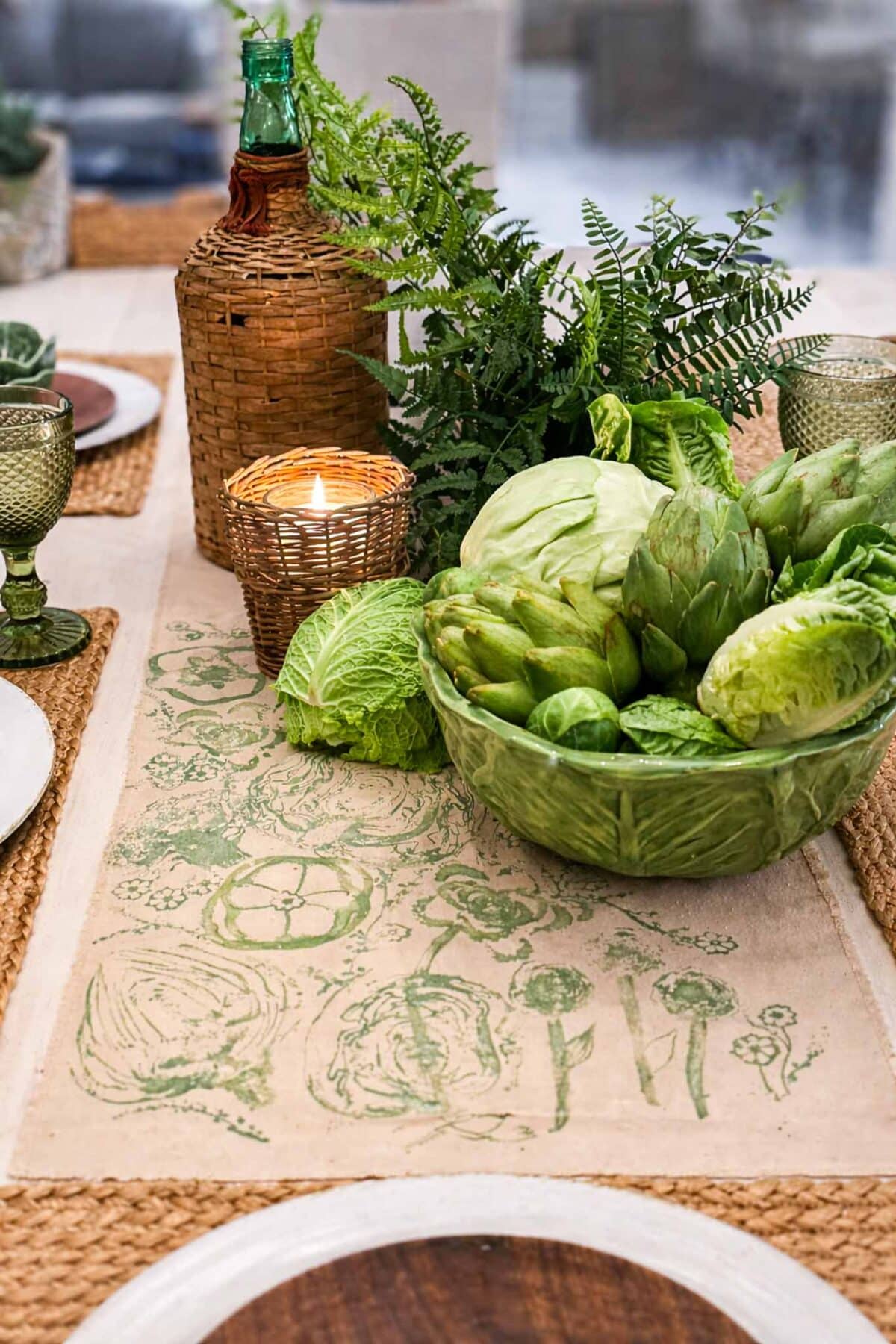 A dining table centerpiece featuring a bowl of green vegetables, a wicker-covered bottle, a lit candle in a wicker holder, and a lush fern arrangement on a botanical-patterned table runner.