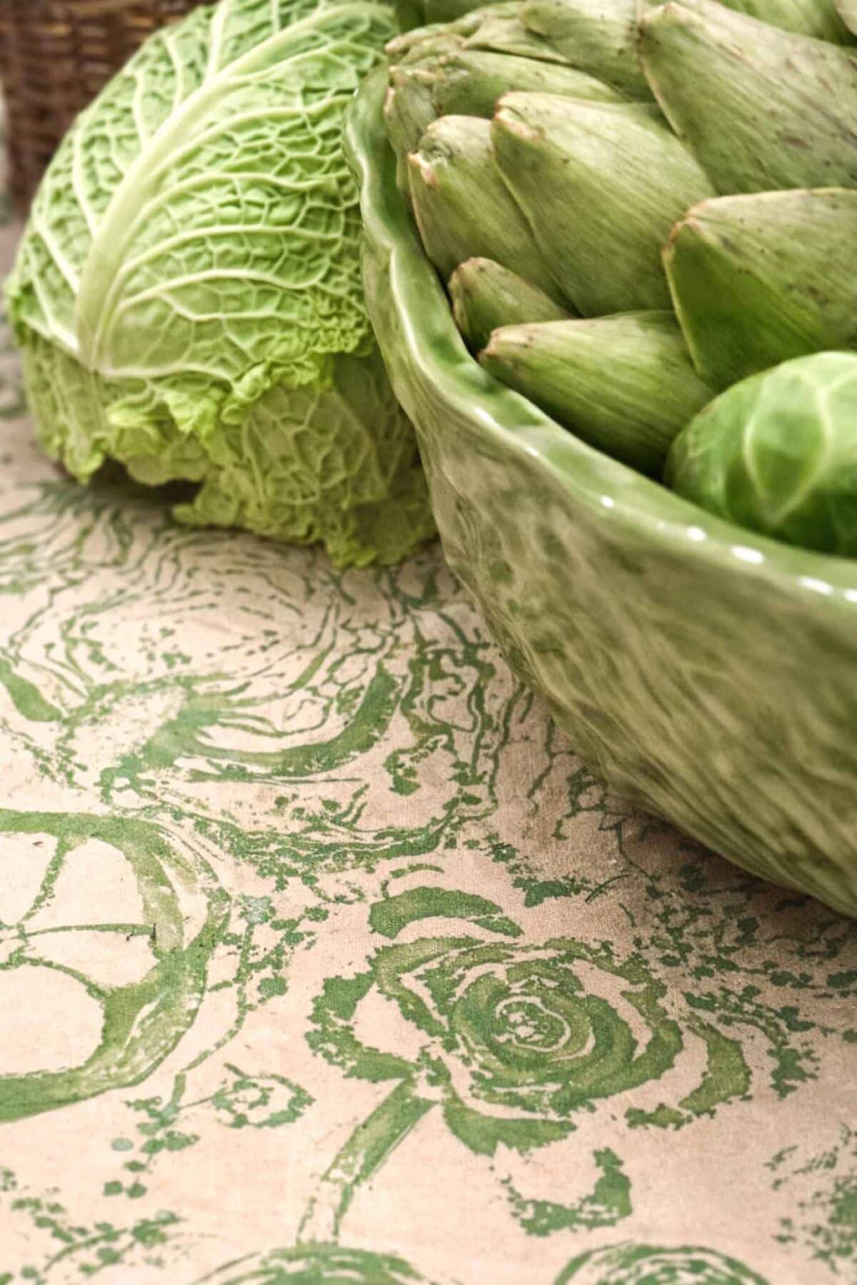 A close-up of a green cabbage and several artichokes in a green ceramic bowl, all resting on a tablecloth with a green floral pattern.