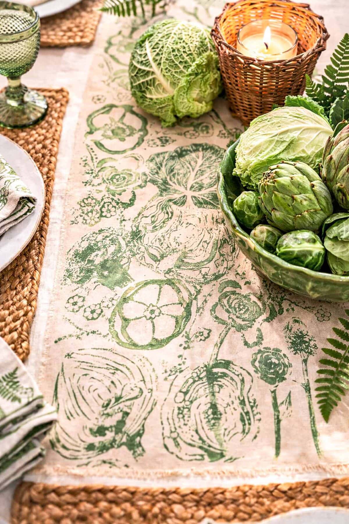 A table set with a linen runner featuring green vegetable prints, surrounded by woven placemats, plates, a green glass goblet, a candle in a wicker holder, a cabbage, and a bowl of artichokes and greens.