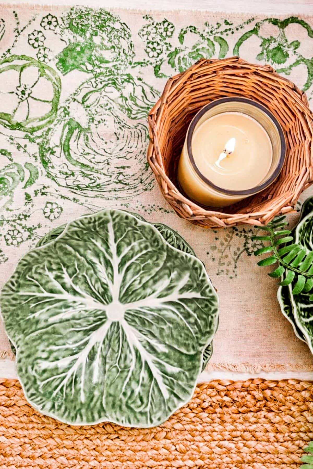 A green leaf-shaped ceramic plate, a wicker basket with a lit candle, and a leafy placemat are arranged on a woven mat, creating a natural, rustic table setting.