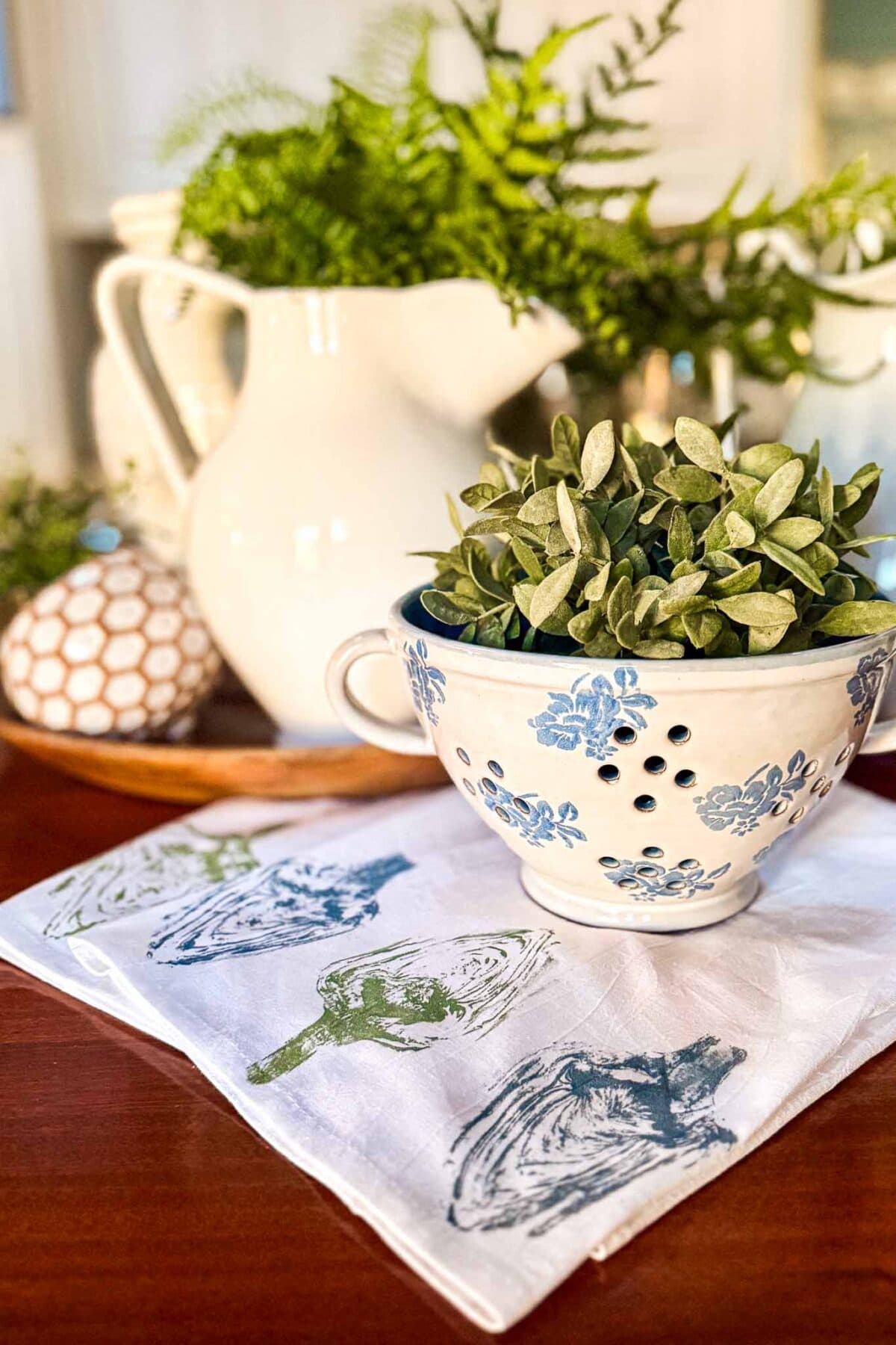 A blue and white floral colander holding a small green plant sits on a napkin with artichoke prints. Behind it is a white pitcher filled with ferns, and a decorative object rests on a wooden tray in the background.