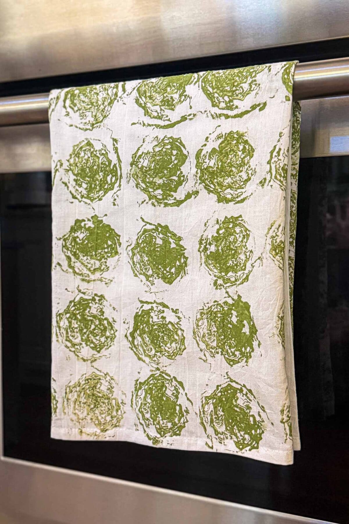 A white kitchen towel with a green circular pattern hangs neatly over a stainless steel oven handle. The pattern appears to be hand-printed, with slight variations in each green circle.