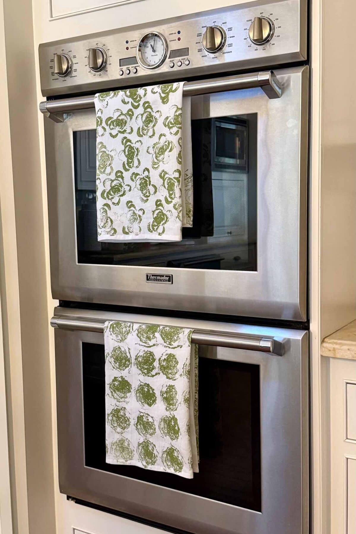 A double wall oven with stainless steel finish, each oven has a towel with a green vegetable print hanging from the handle. The control panel is at the top, with knobs and a clock.