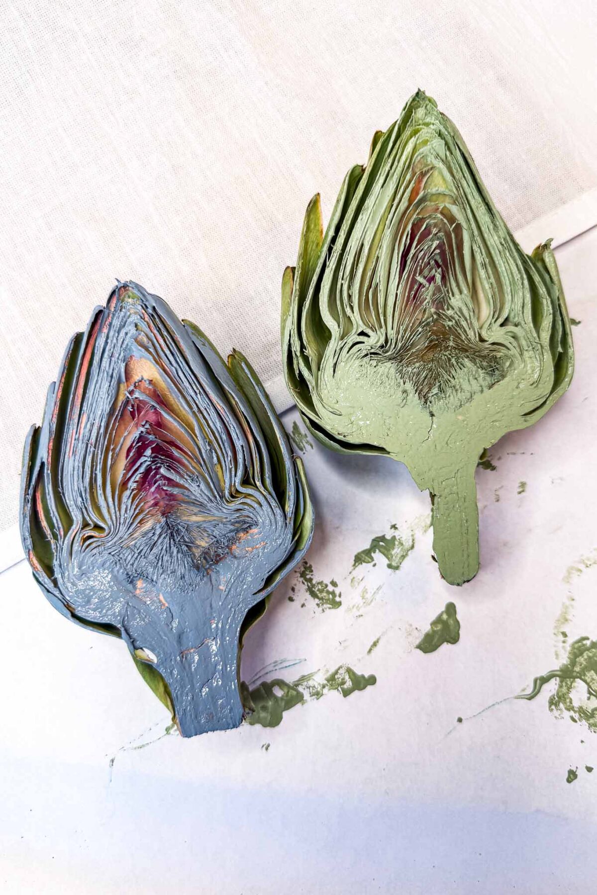 Two artichokes cut in half, one painted green and the other painted blue, lying on a white surface with some paint smudges nearby.