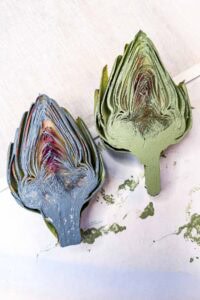 Two artichokes cut in half, one painted green and the other painted blue, lying on a white surface with some paint smudges nearby.
