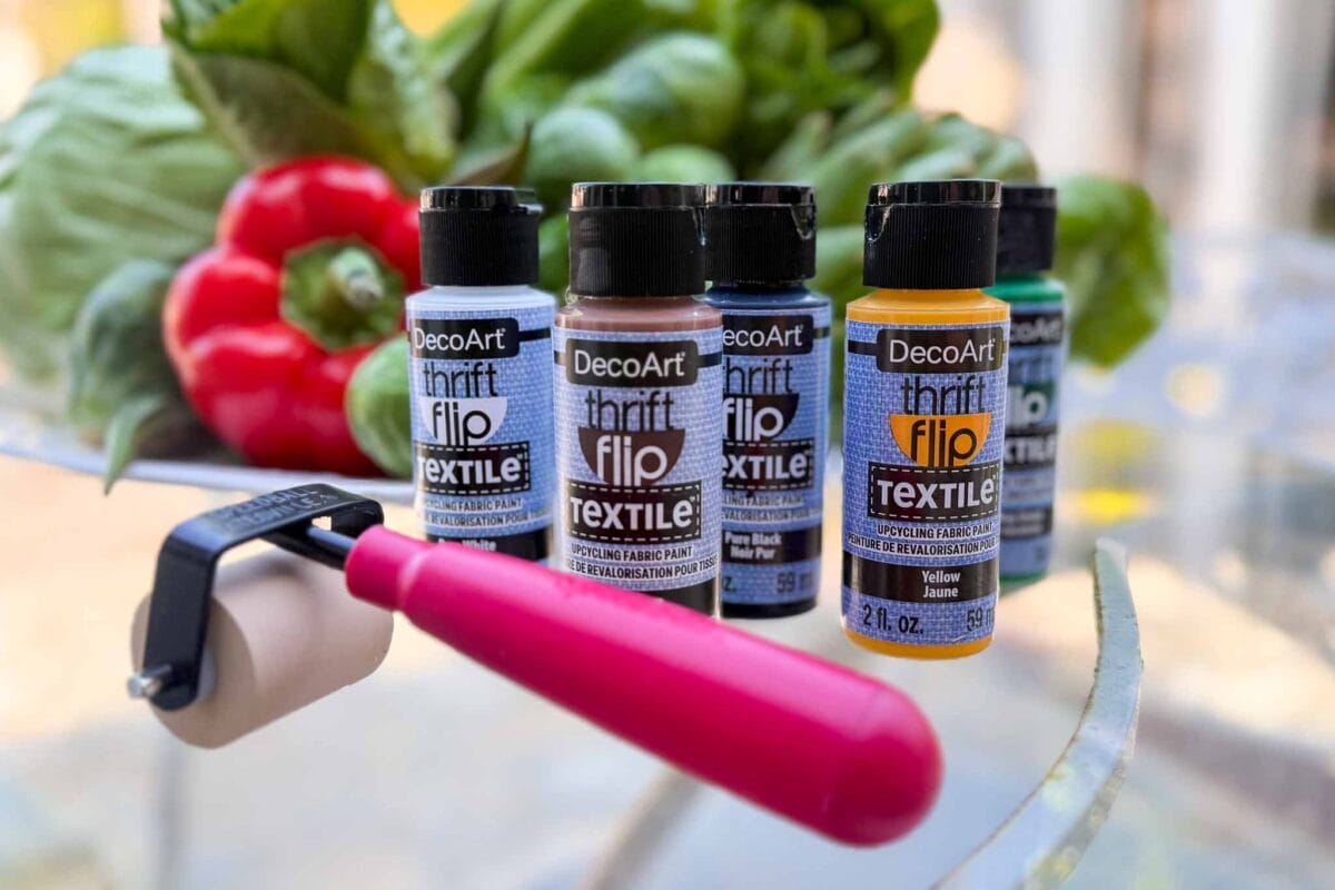 Five bottles of DecoArt Thrift Flip Textile paint stand on a glass table with a small paint roller in front; fresh vegetables, including red peppers and greens, are blurred in the background.