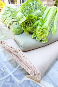 Green vegetables, including cabbage, lettuce, brussels sprouts, and celery, are displayed on two folded fabric pieces—one green and one beige—on a glass surface.