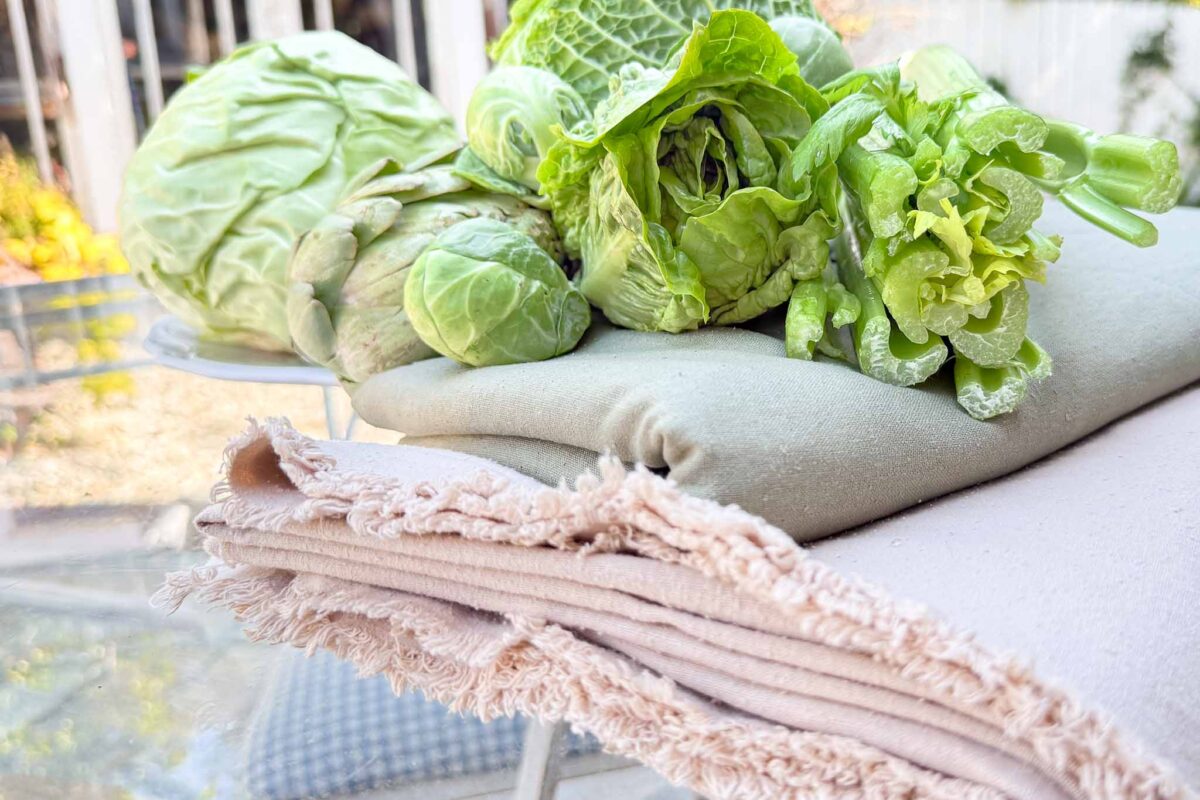 Fresh green vegetables, including cabbage, Brussels sprouts, and lettuce, rest on folded beige and light green fabric with fringed edges, set on a glass table outdoors.