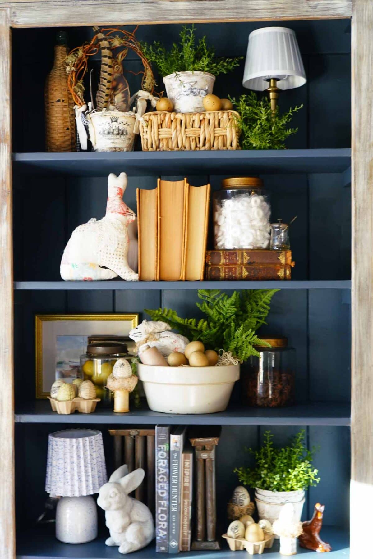 A bookshelf decorated with vintage books, potted greenery, woven baskets, ceramic rabbits, eggs in bowls, glass jars, small lamps, and assorted ornaments arranged on dark blue shelves.