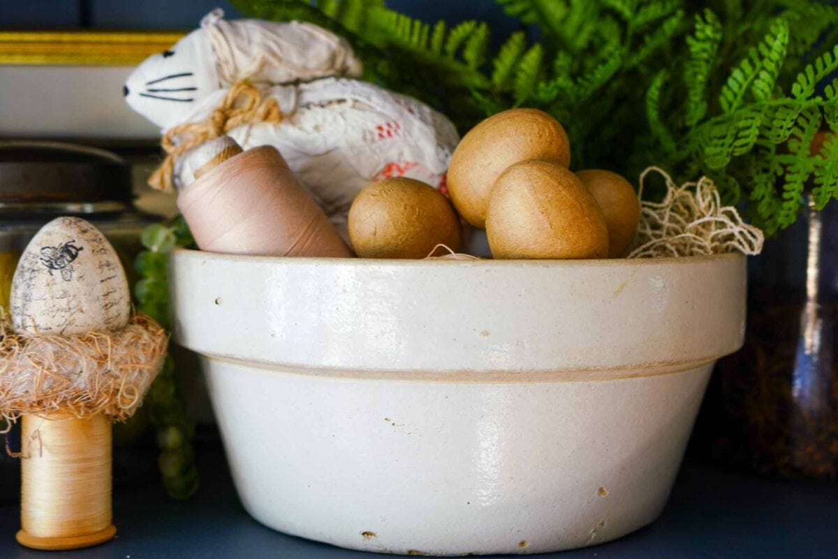 A white ceramic bowl filled with wooden eggs, a spool of thread, a cloth bunny, and decorative netting sits in front of leafy green ferns. Another decorated egg and thread spool are nearby.