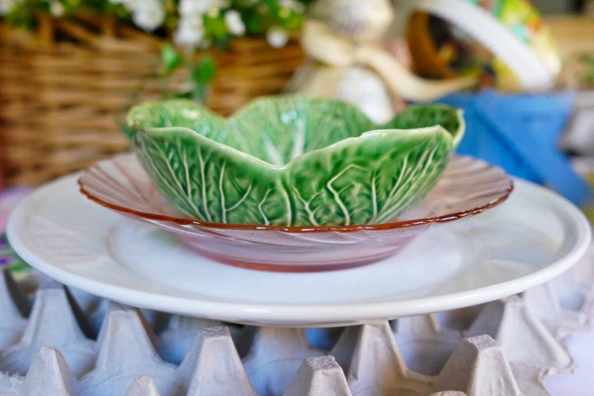 A decorative green bowl shaped like lettuce sits on a pink glass plate, which is placed on a white plate. The plates rest on an egg carton, with a wicker basket and flowers in the blurred background.