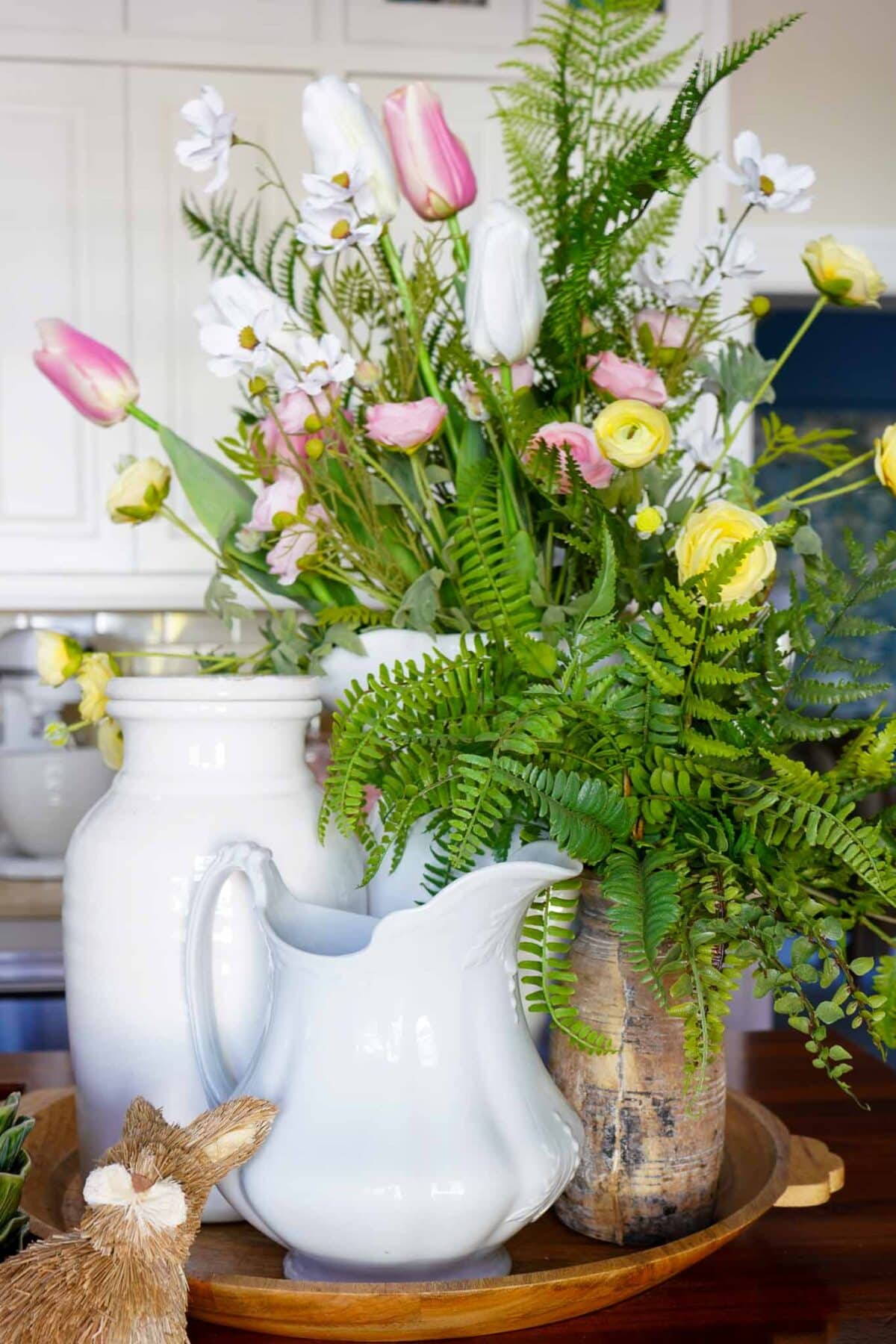 A spring-themed arrangement with pink, yellow, and white flowers, green ferns in vases, a white ceramic pitcher, and a small decorative bunny on a wooden tray, set on a kitchen counter.