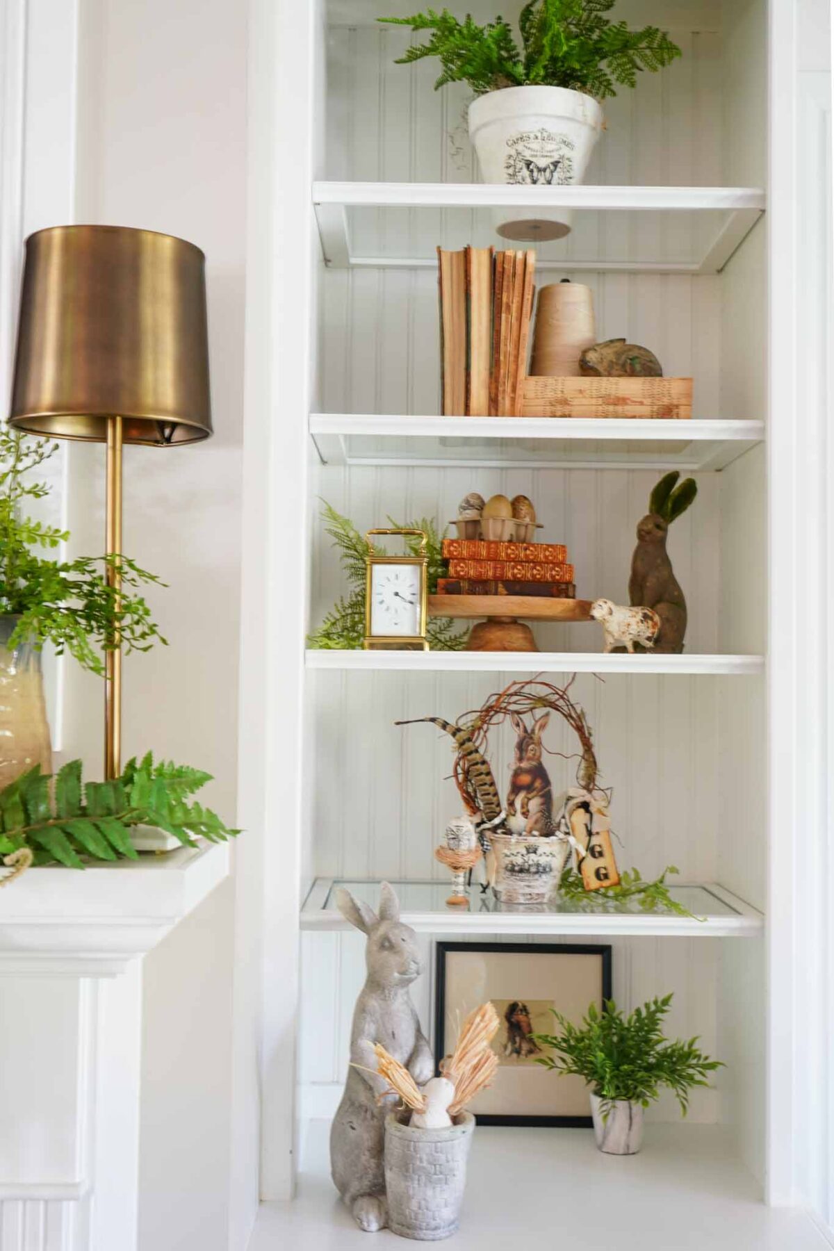 White built-in shelves display ferns, vintage books, wooden eggs, a gold clock, ceramic rabbits, a lamp with a gold shade, and framed art, creating a cozy and decorative arrangement.