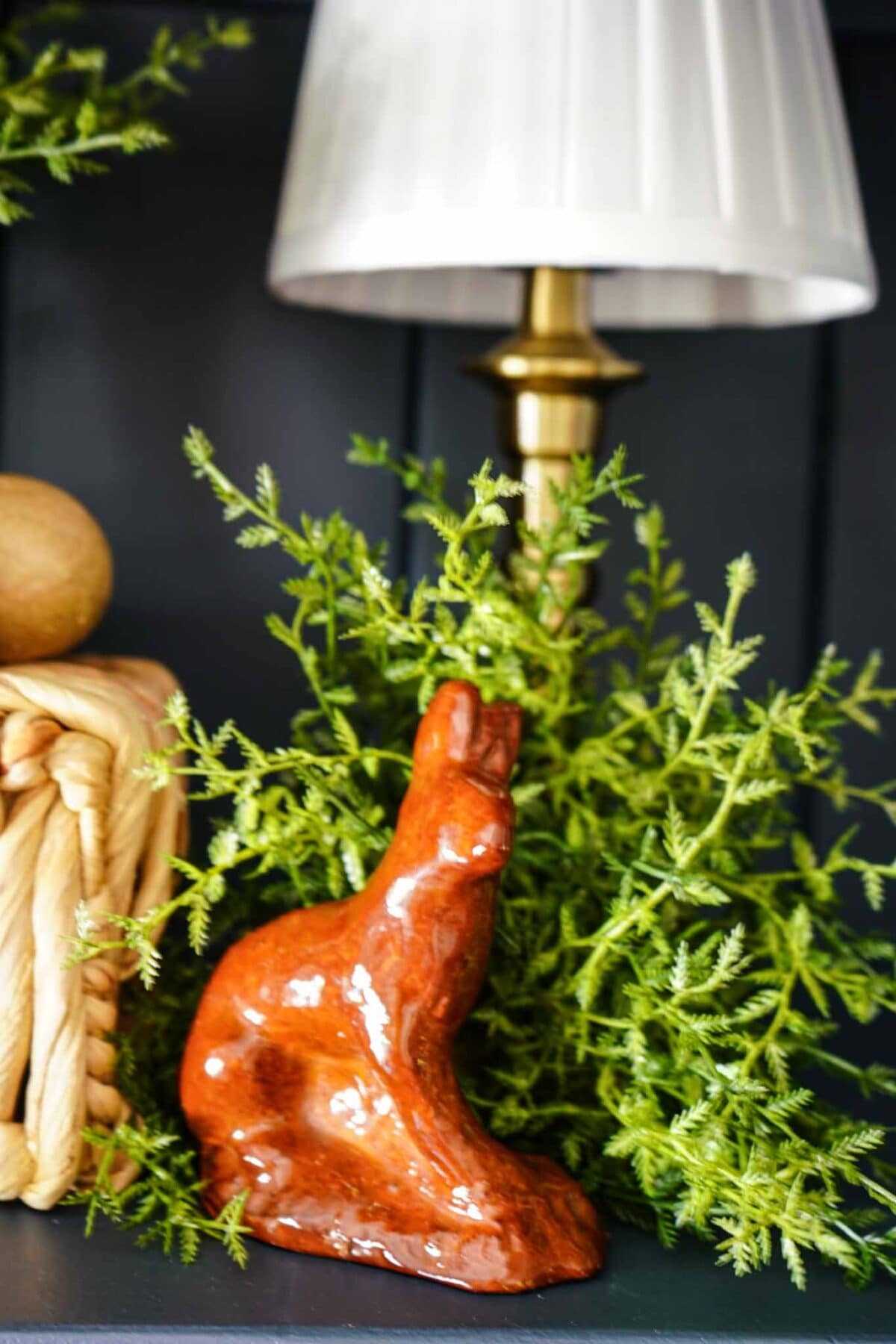 A glossy orange ceramic rabbit figurine sits in front of green foliage, beside a gold lamp with a white pleated shade, and a coil of thick rope on a dark shelf.