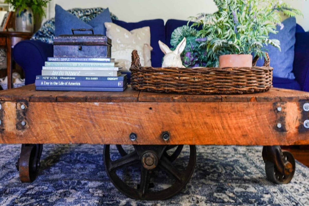A rustic wooden coffee table with metal wheels holds a stack of books, a metal box, a woven basket with a white rabbit figurine, and a potted plant. A blue sofa with patterned pillows is in the background.