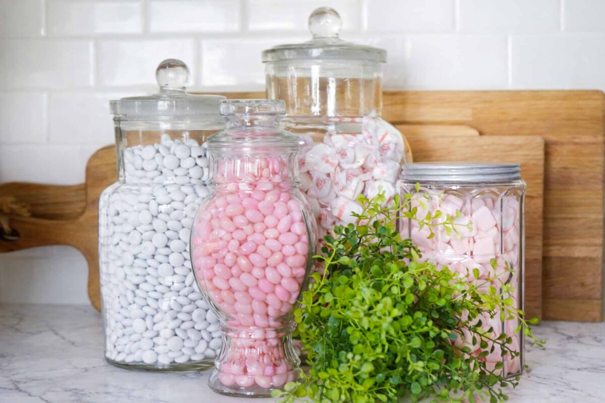 Three glass jars filled with white, pink, and red candies are arranged on a marble countertop, with a small green potted plant in front. Wooden cutting boards lean against a white tile backsplash in the background.