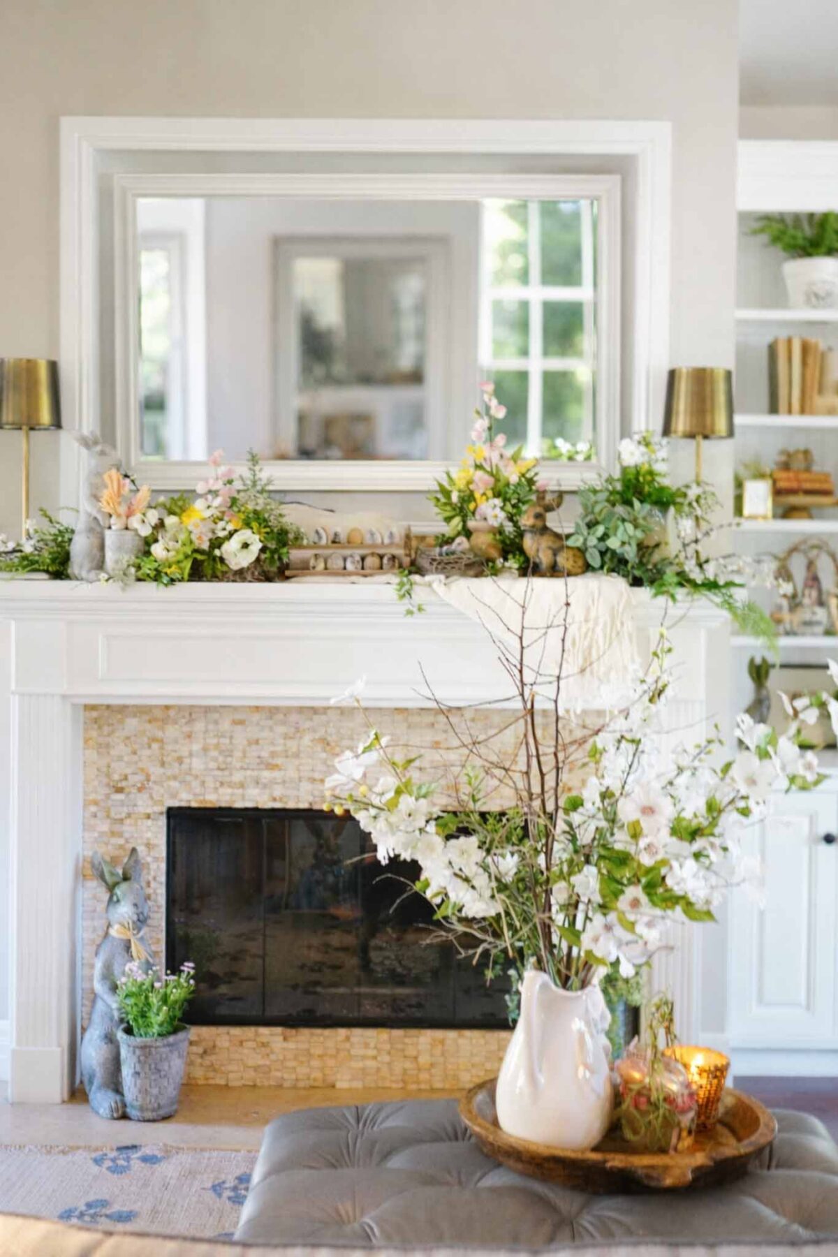A bright living room features a decorated white fireplace with flowers, greenery, candles, and a large mirror above. A vase with blooming branches and a tray with glass jars sit on a nearby ottoman.