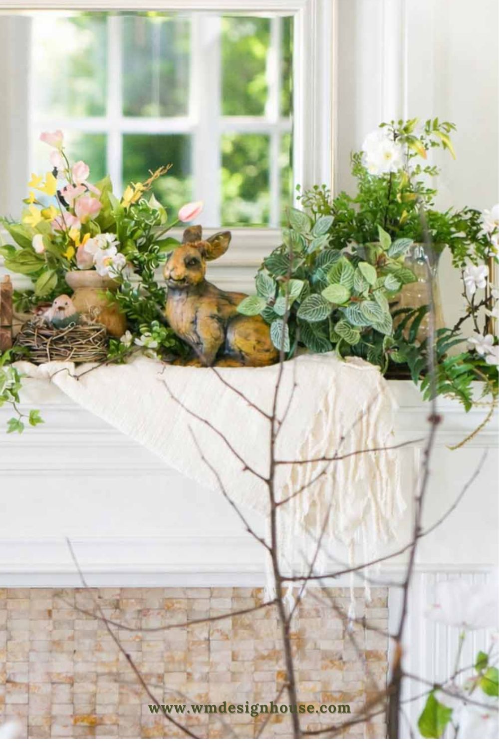 A mantel decorated with spring flowers, green plants, a bird’s nest, and a small deer figurine offers charming vintage Easter decorating ideas. A cream-colored cloth is draped underneath, with a window reflecting greenery in the background.