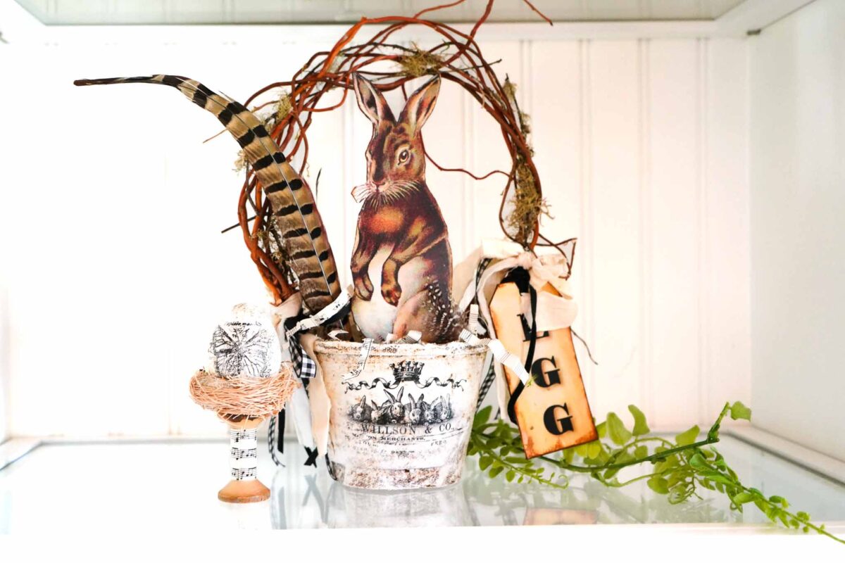 A decorative arrangement featuring a rabbit cutout in a vintage-style pot with feathers, twigs, and "EGG" tags on a white shelf with greenery—perfect for those seeking charming vintage Easter decorating ideas.
